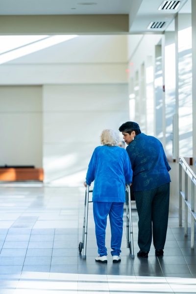A nurse assists an elderly woman with a walker in a bright hospital corridor.